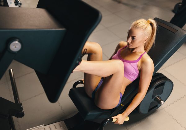 Young pretty blond woman, with pony tale, pink top and mini shorts, working her legs at the machine press in the gym. Top angle shot.